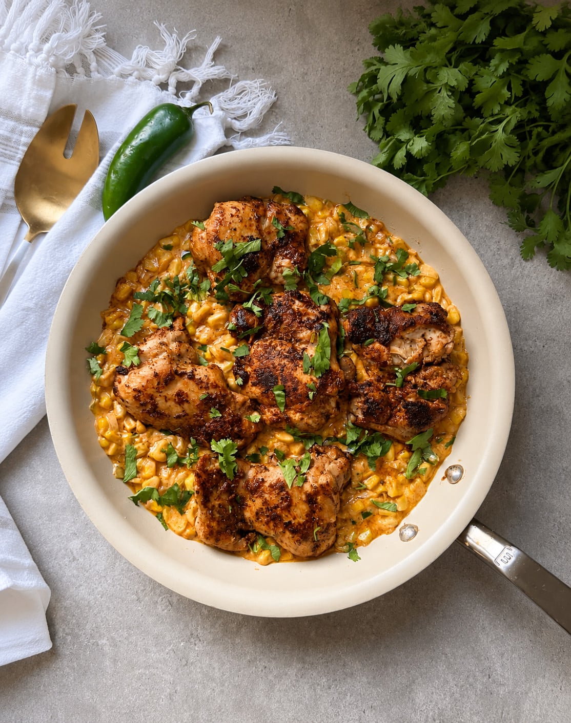 A skillet with seasoned chicken thighs over creamy corn, garnished with chopped cilantro&mdash;this Chicken Thighs with Jalape&ntilde;o Creamed Corn recipe makes the perfect one-pan dinner. Fresh cilantro, a jalape&ntilde;o, and utensils are nearby on a gray surface.