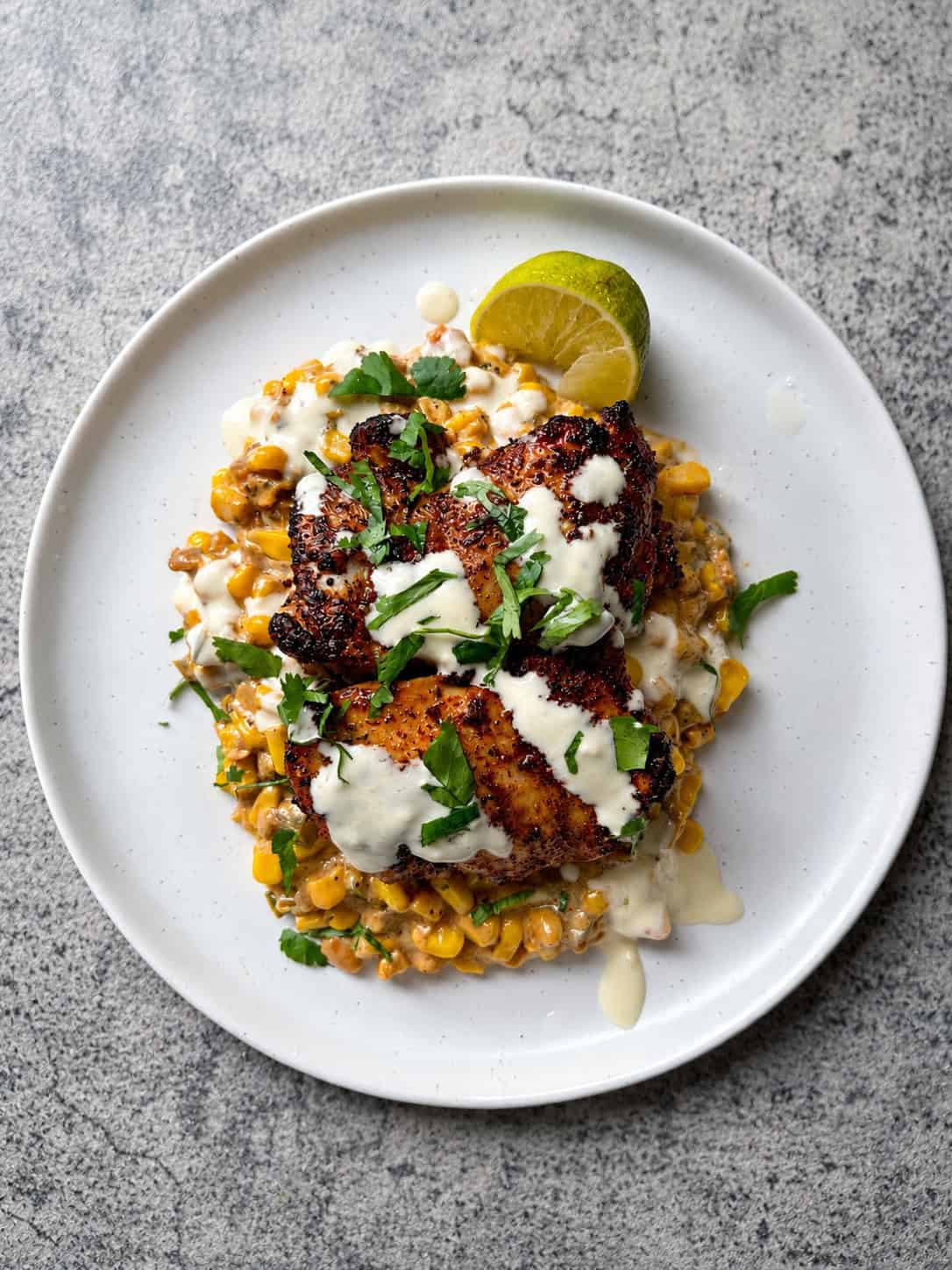 A white plate with grilled chicken thighs on a bed of jalapeño creamed corn, topped with creamy white sauce and chopped herbs, garnished with a lime wedge—an easy one-pan dinner served on a gray surface.