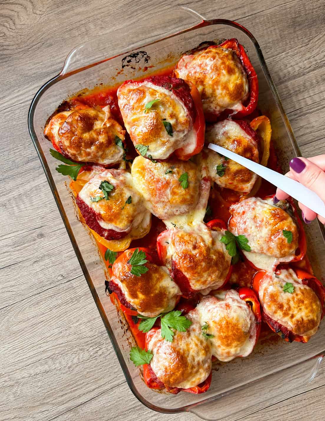 A glass baking dish filled with stuffed bell peppers topped with melted cheese and herbs. A hand is using a white spatula to lift one pepper. The dish sits on a wooden surface.