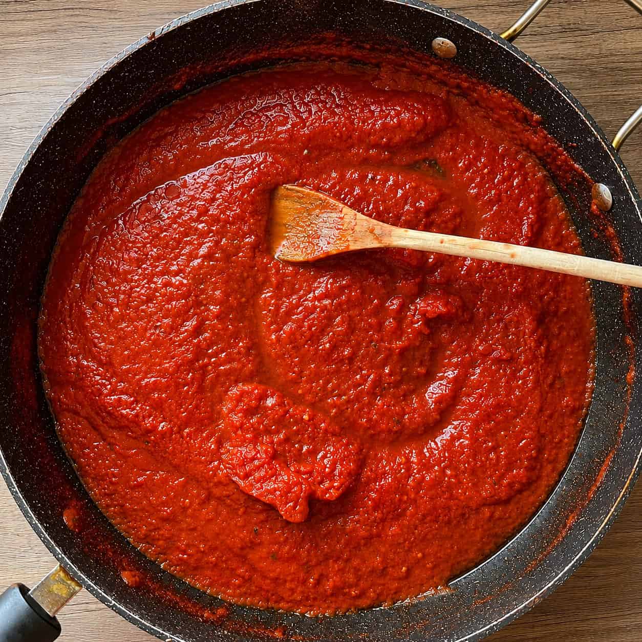 A top-down view of a speckled black non-stick pan filled with thick, vibrant red tomato sauce. A light-colored wooden spoon rests horizontally in the center of the sauce, which is being cooked on a light wood-grained surface.