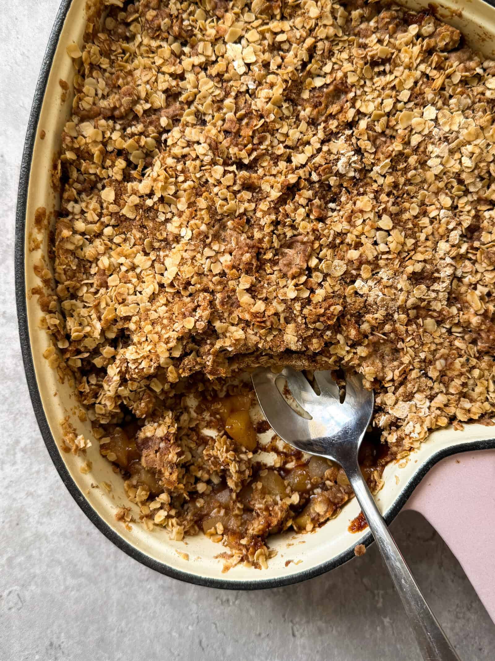 A close-up of a baked fruit crumble in a large dish, topped with a golden oat and brown sugar crust. A portion has been scooped out with a large metal spoon, revealing the fruit filling—perfect for an Easy Apple Crisp Recipe.