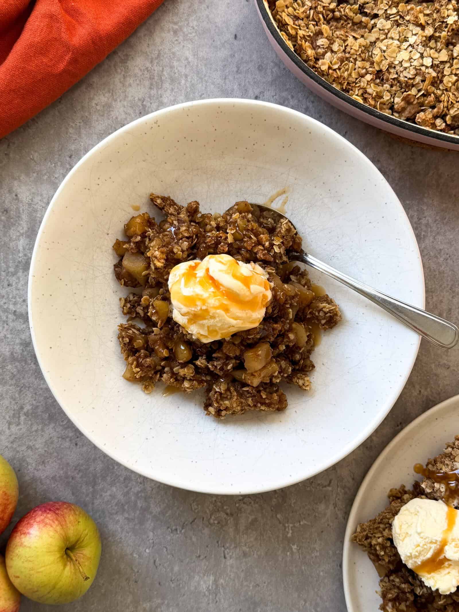 A plate of apple crisp topped with vanilla ice cream and caramel drizzle sits on a gray surface, with a spoon in the bowl. Fresh apples and a pan of more apple crisp suggest this is the Best Apple Crisp Recipe for any occasion.