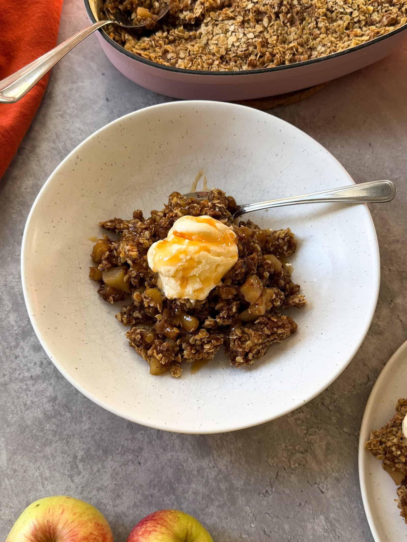 A bowl of apple crisp topped with vanilla ice cream and caramel drizzle, with a spoon in the bowl. A baking dish of Easy Apple Crisp and fresh apples rest nearby on a gray surface.