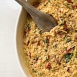 A white bowl filled with creamy orzo pasta mixed with sun-dried tomatoes, spinach, and herbs. A wooden spoon rests in the bowl, showcasing the colorful ingredients and texture of the dish.