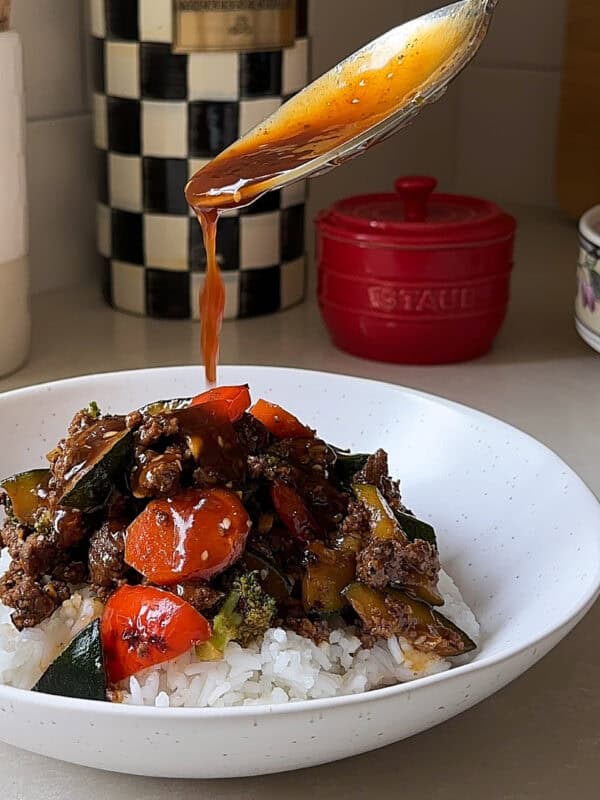 A spoon drizzles sauce over a bowl of rice topped with colorful stir-fried vegetables and savory ground beef, creating a delicious Ground Beef Stir Fry. In the background are a checkered container, a red dish, and kitchen utensils.