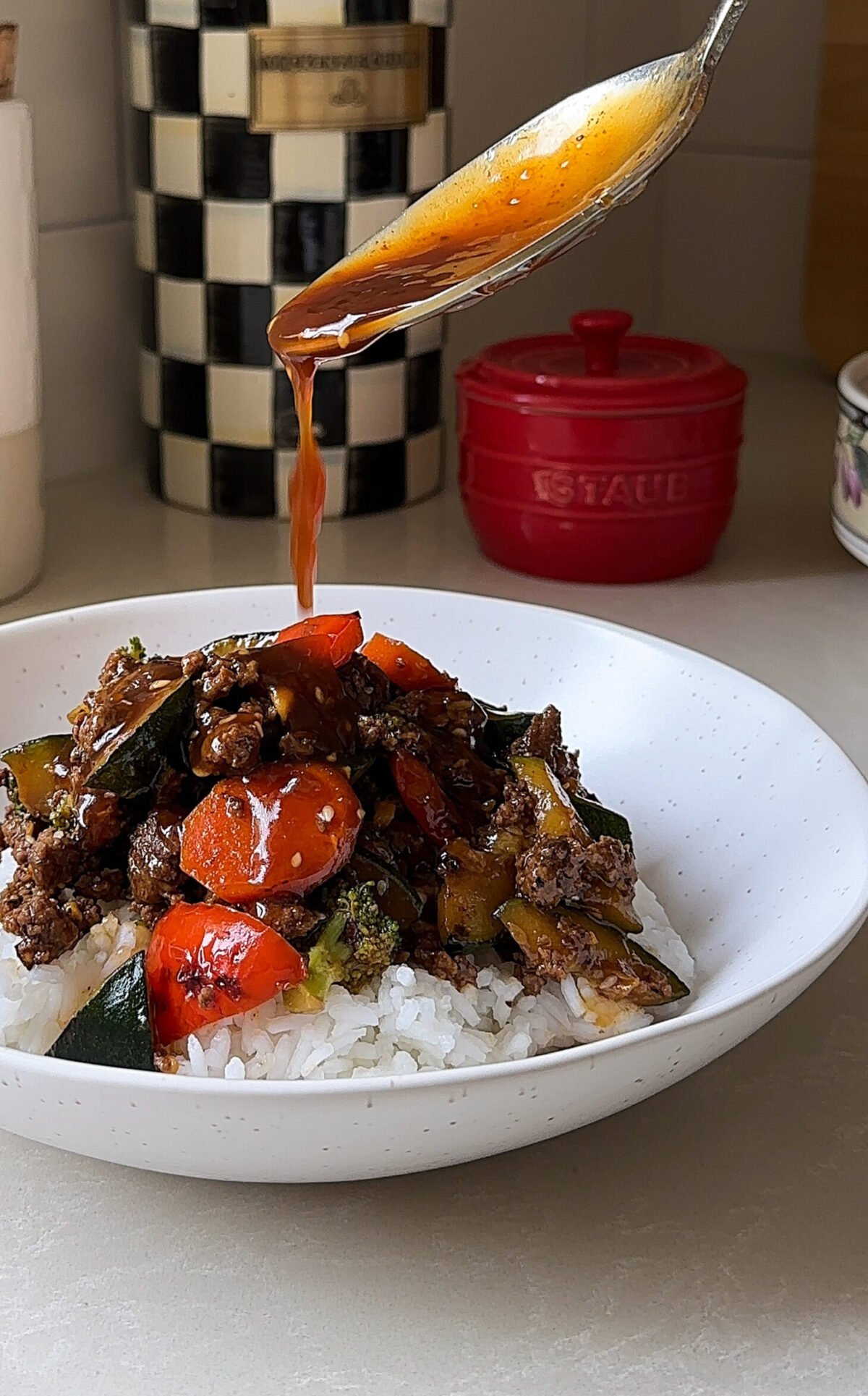 A spoon drizzles sauce over a bowl of rice topped with stir-fried beef and vegetables, showcasing a delicious Ground Beef Stir Fry Recipe with carrots, zucchini, and broccoli in a kitchen setting.
