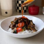 A spoon drizzles sauce over a bowl of white rice topped with a colorful Ground Beef Stir Fry, featuring carrots, zucchini, and bell peppers, on a kitchen counter with jars and containers in the background.