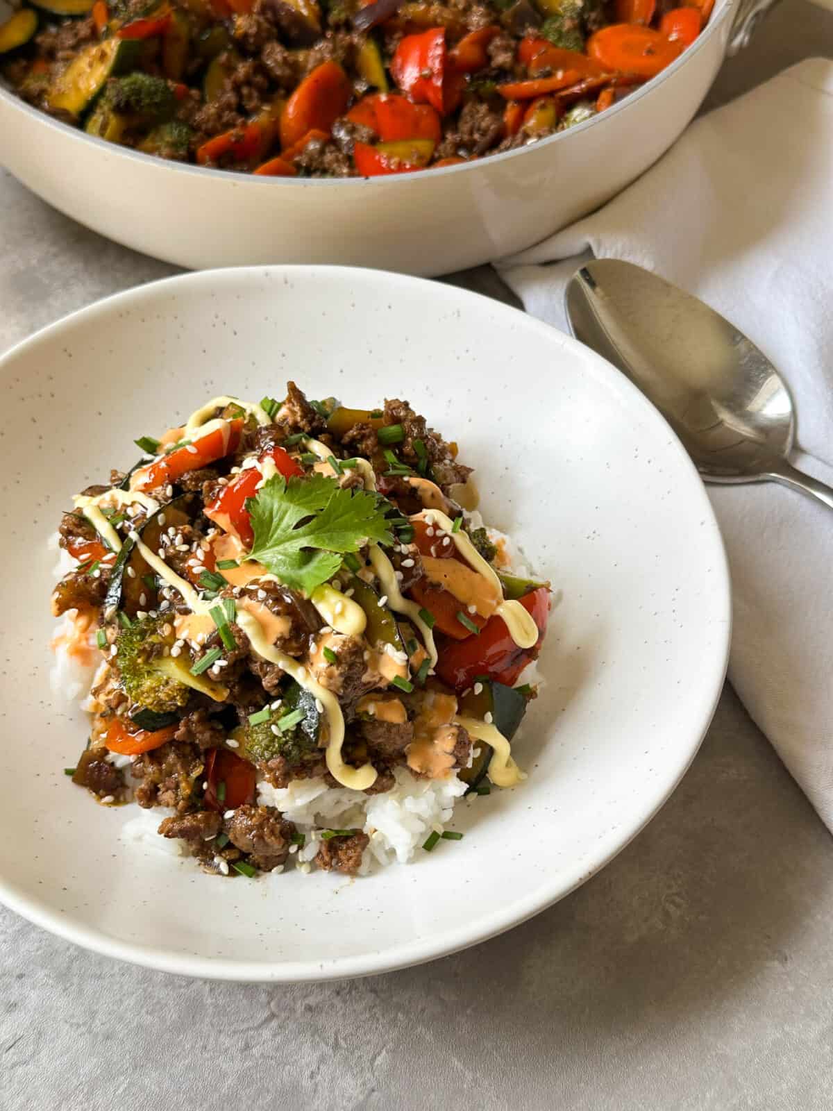 A bowl of white rice topped with stir-fried vegetables and ground beef, drizzled with creamy sauce and garnished with sesame seeds and cilantro. This Ground Beef Stir Fry Recipe is served with a spoon, napkin, and skillet of more stir-fry in the background.