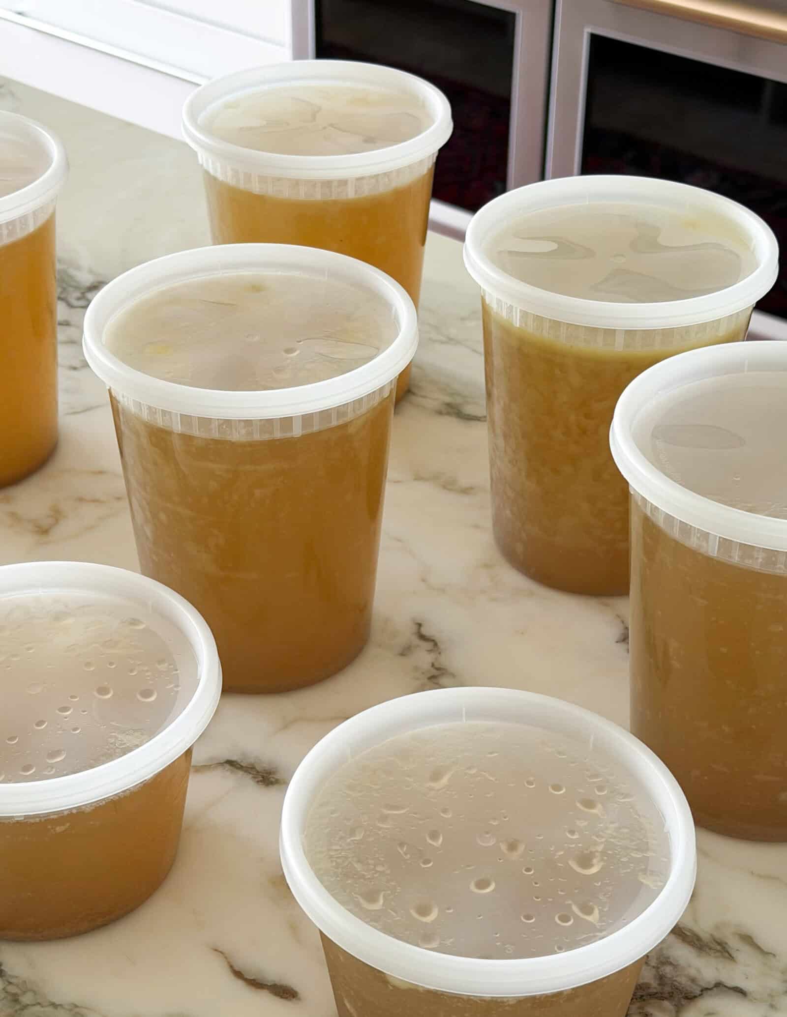 Several clear plastic containers filled with homemade chicken broth, sealed with lids, are arranged on a marble kitchen countertop. Ovens are visible in the background.