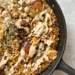 A skillet filled with baked rice and browned chicken pieces, garnished with a drizzle of creamy sauce and sprigs of fresh herbs. The crispy texture of the rice and chicken is visible, creating an appetizing appearance.