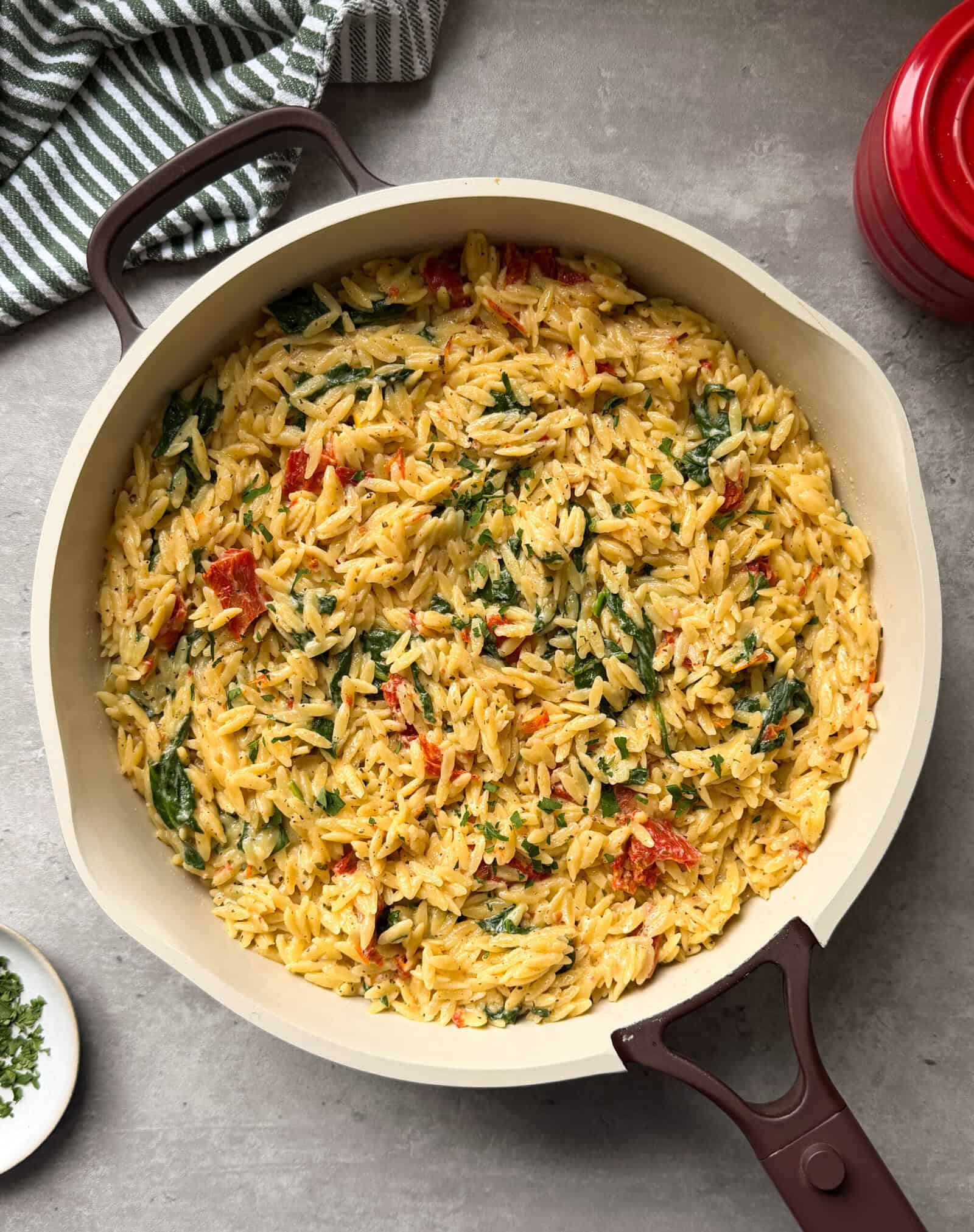 A large skillet filled with creamy Boursin Spinach and Sun-dried Tomato Orzo Bake sits on a gray surface, with a striped towel and a red pot partially visible nearby—perfect for fans of comforting orzo recipes.