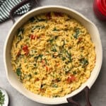 A large skillet filled with Boursin Spinach and Sun-dried Tomato Orzo Bake sits on a gray countertop. Creamy orzo pasta is mixed with spinach, sun-dried tomatoes, and herbs, with a striped kitchen towel and a red lid nearby.