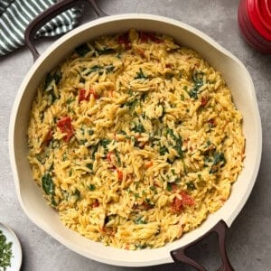 A large skillet filled with Boursin Spinach and Sun-dried Tomato Orzo Bake sits on a gray countertop. Creamy orzo pasta is mixed with spinach, sun-dried tomatoes, and herbs, with a striped kitchen towel and a red lid nearby.