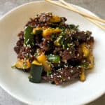 A dish of stir-fried beef and zucchini garnished with sesame seeds and chopped green onions is served on a white plate. A pair of wooden chopsticks rests on the plates rim.
