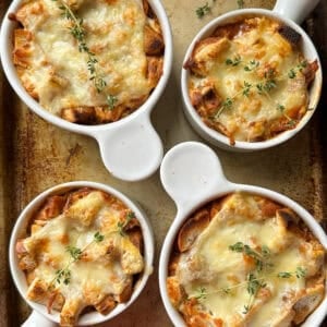 Four white ramekins filled with an easy French Onion Soup recipe, topped with melted cheese and garnished with fresh thyme, sit on a rustic baking sheet.
