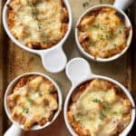 Four round baking dishes filled with French onion soup, topped with toasted bread and melted cheese. Fresh thyme sprigs garnish the soups, which are placed on a rustic metal baking tray.