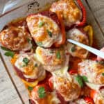 A glass baking dish filled with stuffed bell peppers topped with melted cheese and tomato sauce. The peppers are garnished with fresh parsley. A white spatula is lifting one pepper from the dish. The dish is on a wooden surface.
