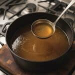 A metal ladle lifting golden brown homemade chicken broth from a black cast iron pot on a wooden cutting board, with a stove visible in the background.
