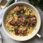 A white skillet filled with seasoned grilled chicken pieces rests on a bed of Jalapeño Creamed Corn, garnished with chopped cilantro. A green pepper and fresh cilantro sit nearby, with a white cloth and a golden serving fork ready to complete this one-pan dinner masterpiece.