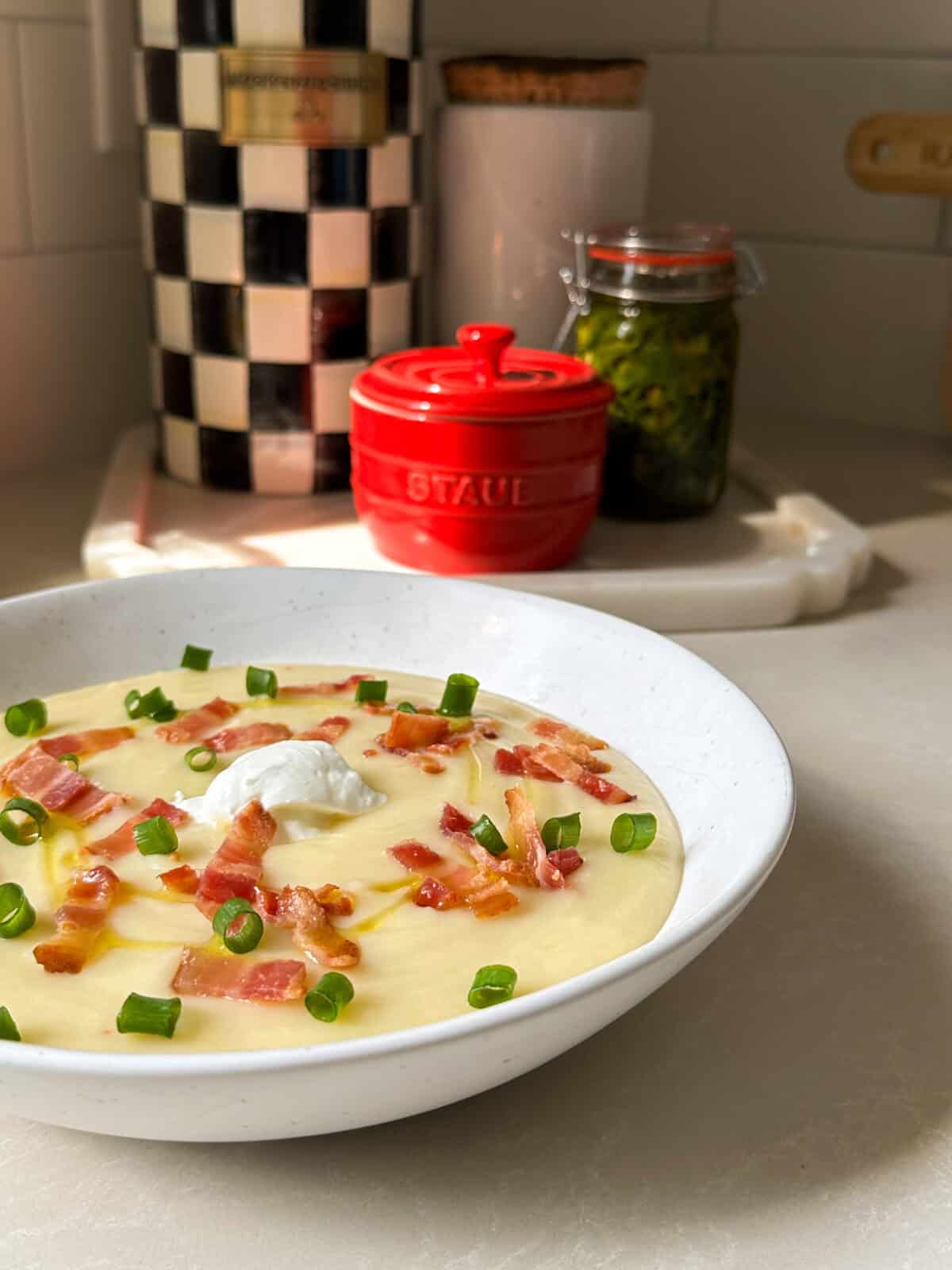 A bowl of creamy potato and leek soup topped with bacon bits, chopped green onions, and a dollop of sour cream sits on a kitchen counter, with kitchen containers and a jar in the background.