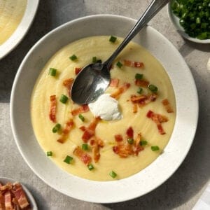 A bowl of creamy potato and leek soup topped with crispy bacon bits, chopped green onions, and a dollop of sour cream. A spoon rests in the bowl, with extra toppings nearby—perfect inspiration if you’re wondering how to make potato soup at home.