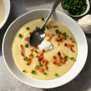A bowl of creamy potato and leek soup topped with bacon bits, chopped green onions, and a dollop of sour cream. A spoon rests in the bowl, with small bowls of green onions and bacon pieces nearby on a gray surface.