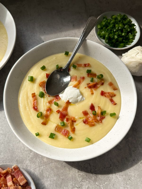 A bowl of creamy potato and leek soup topped with bacon bits, chopped green onions, and a dollop of sour cream. A spoon rests in the bowl, with small bowls of green onions and bacon pieces nearby on a gray surface.