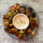 A gray plate of crispy, golden-brown Brussels sprouts surrounds a white bowl filled with a creamy dipping sauce, garnished with chopped herbs, on a textured stone surface.