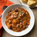 A bowl of beef stew with chunks of meat, potatoes, carrots, and herbs in a rich brown sauce, served on a wooden table with cheddar biscuits on a plate and an orange napkin beside a spoon.