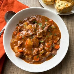 A bowl of beef stew with chunks of meat, potatoes, carrots, and herbs in a rich brown sauce, served on a wooden table with cheddar biscuits on a plate and an orange napkin beside a spoon.