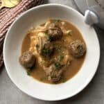 A white plate holds meatballs and mashed potatoes, topped with brown gravy and garnished with chopped herbs. A patterned cloth and the edge of a slow cooker are visible beside the plate on a gray surface.