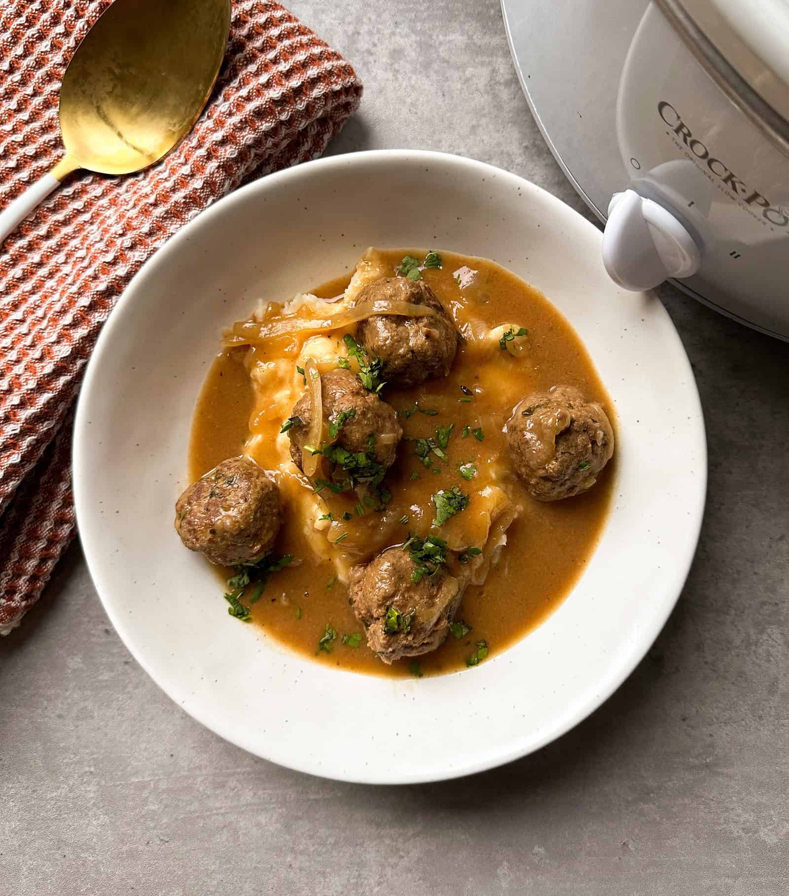 A white plate with meatballs in brown gravy, garnished with herbs, served over mashed potatoes. A crockpot, spoon, and folded textured napkin are nearby on a light-colored surface.