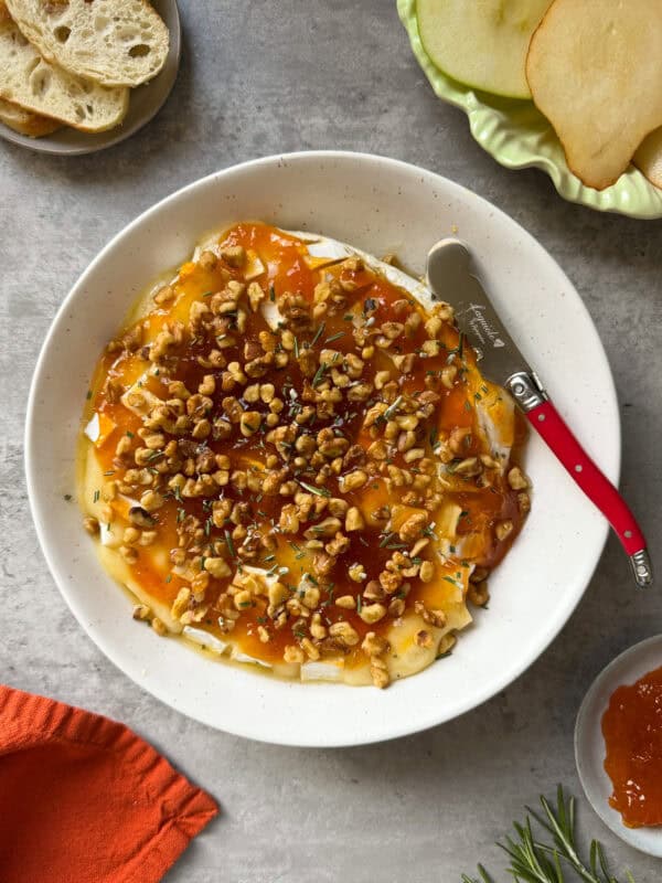 A round wheel of brie cheese, perfect for a Baked Brie Appetizer, is topped with chopped walnuts and apricot preserves on a white plate. A red-handled cheese knife awaits nearby, with slices of bread and apples arranged on a light gray surface.