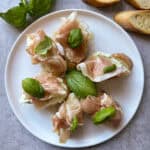A plate of appetizers featuring slices of prosciutto, fresh basil leaves, and green toppings on bread slices with a creamy spread. Two slices of bread are set beside the plate. The background is a light gray surface.