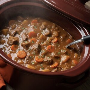A hearty Crockpot Beef Stew Recipe with chunks of beef, carrots, and potatoes in a thick broth, served in a red slow cooker with a ladle. Fresh biscuits can be seen on a plate in the background.