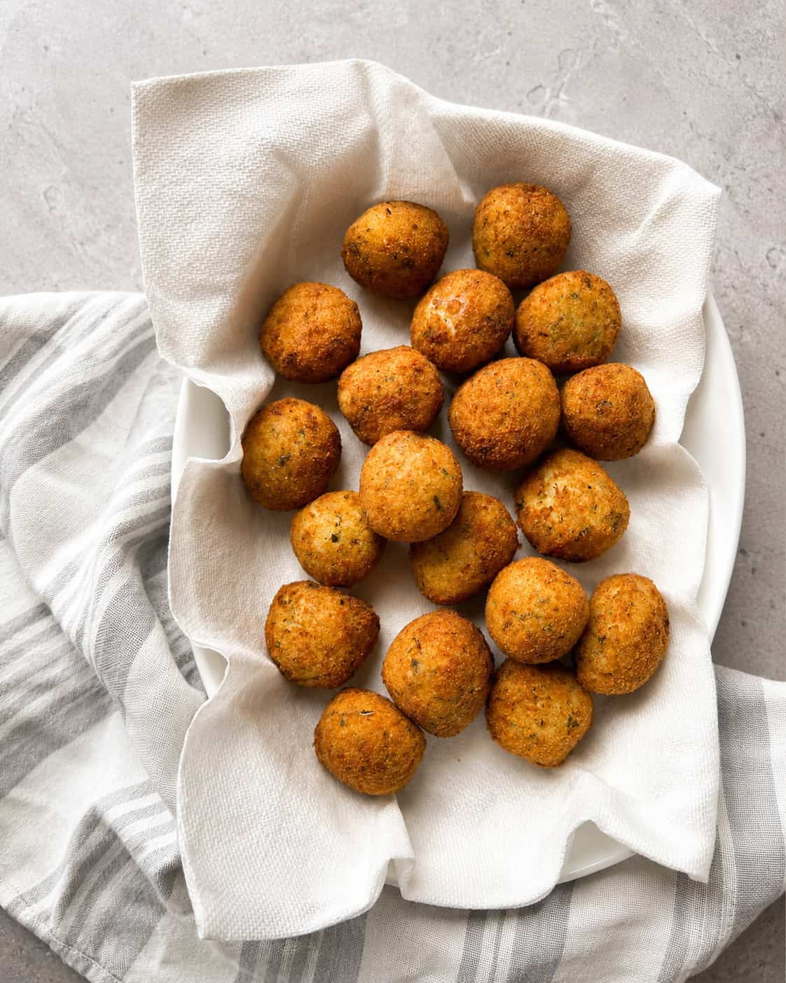 A white bowl lined with a paper towel holds golden-brown falafel balls, placed on a gray surface with a striped white and gray cloth underneath.
