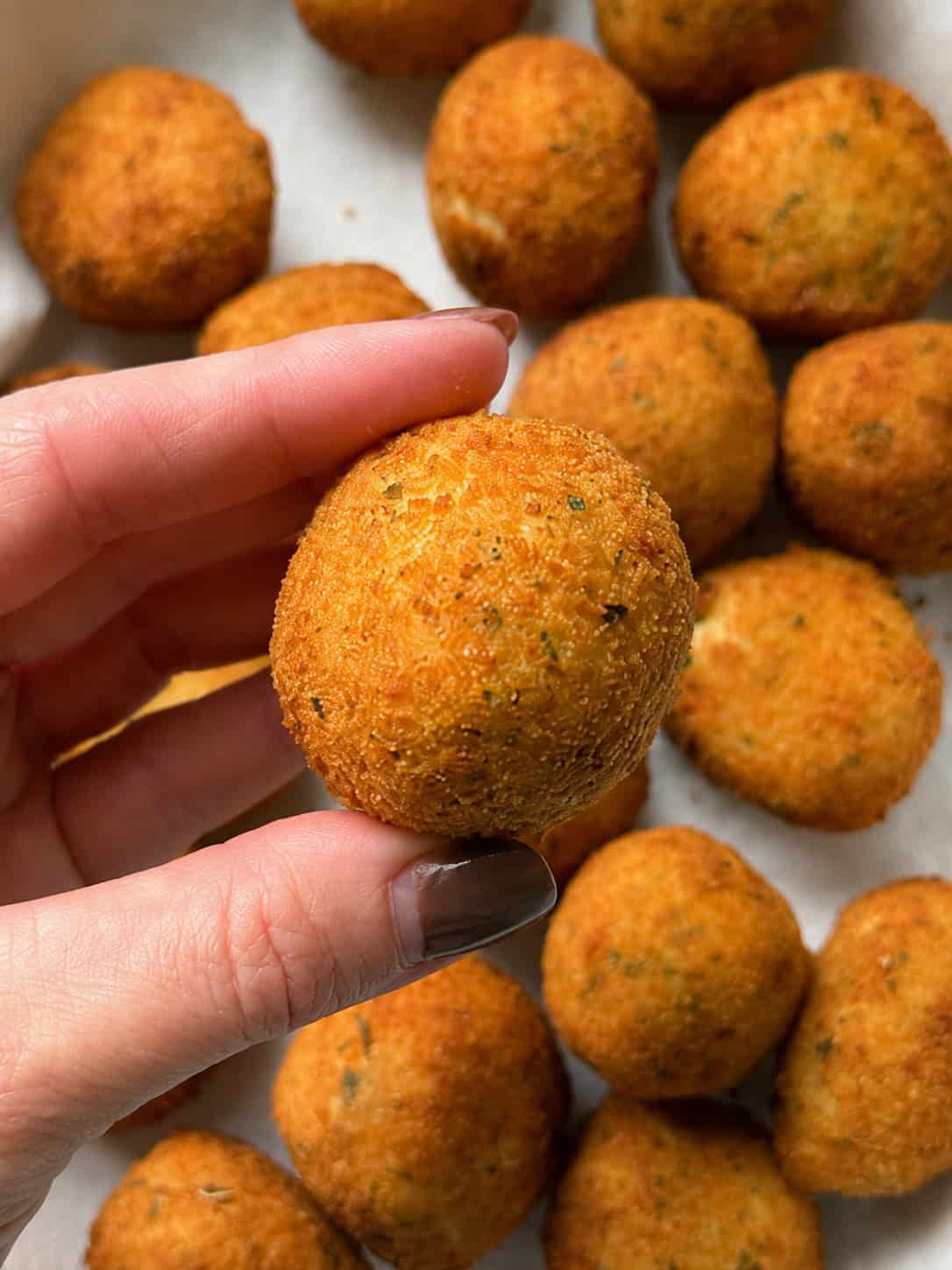 A hand holding a round, golden-brown fried snack with a textured surface, with several more similar snacks in the background.