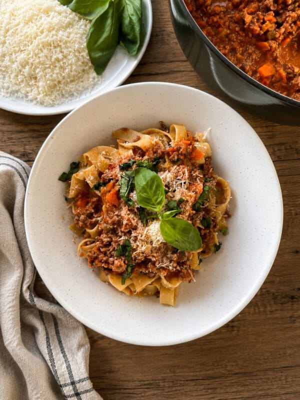 A plate of tagliatelle pasta topped with meat sauce, grated cheese, and basil leaves sits on a wooden table beside a bowl of grated cheese, fresh basil, a pot of meat sauce, and a striped cloth napkin.