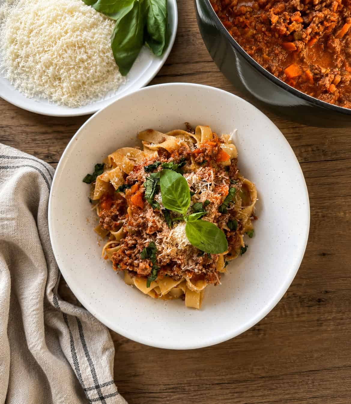 A plate of tagliatelle pasta topped with meat sauce, grated cheese, and basil leaves sits on a wooden table beside a bowl of grated cheese, fresh basil, a pot of meat sauce, and a striped cloth napkin.