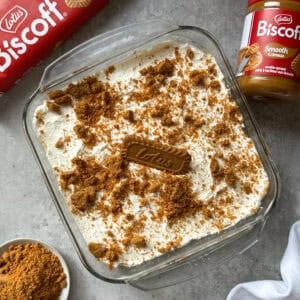 A glass dish of creamy Biscoff No Bake Cheesecake topped with crushed Lotus Biscoff cookies, surrounded by a pack of biscuits, a jar of Biscoff spread, and a bowl of crushed cookies on a gray surface.