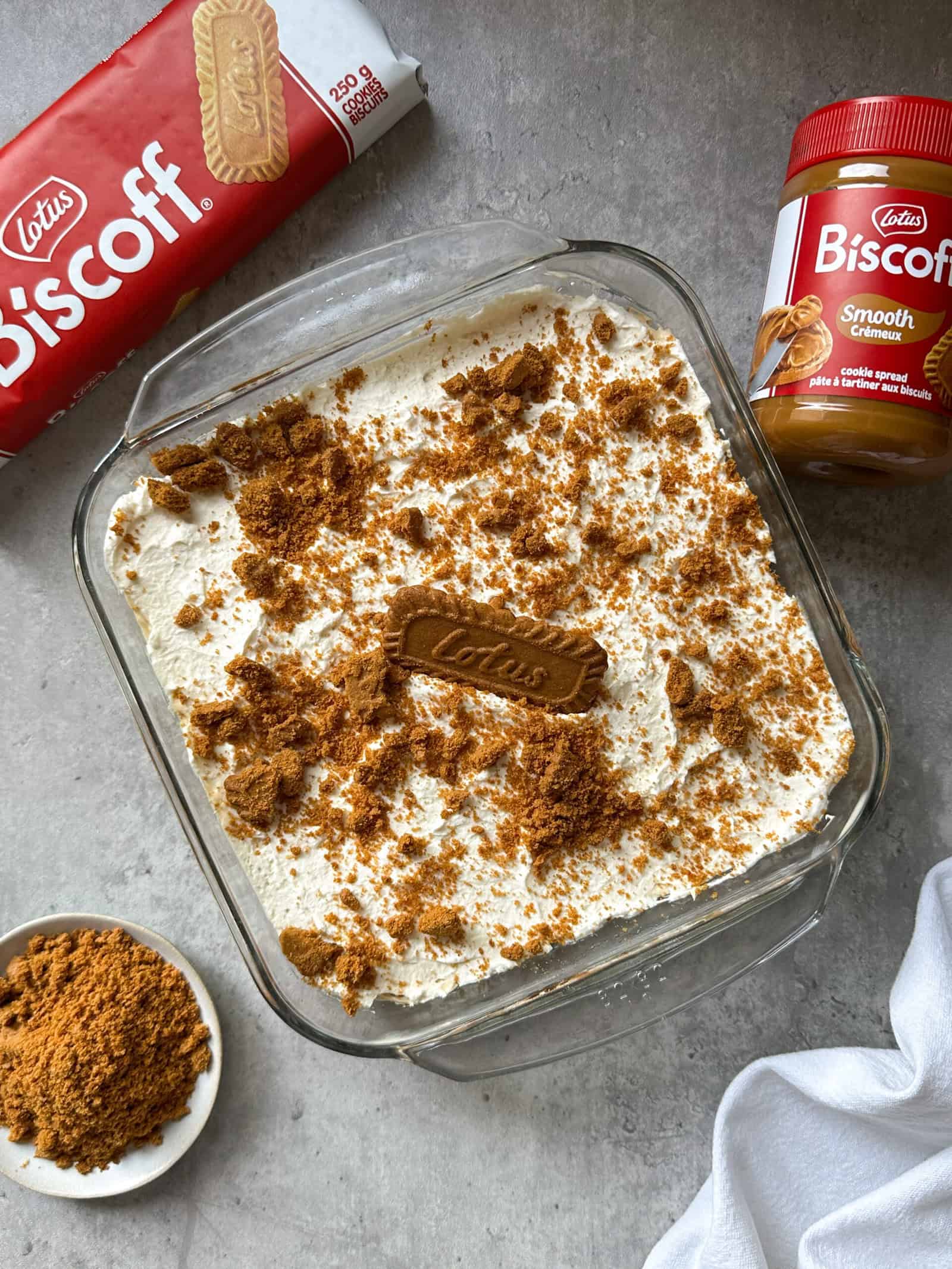 A glass dish of creamy Biscoff No Bake Cheesecake topped with crushed Lotus Biscoff cookies, surrounded by a pack of biscuits, a jar of Biscoff spread, and a bowl of crushed cookies on a gray surface.