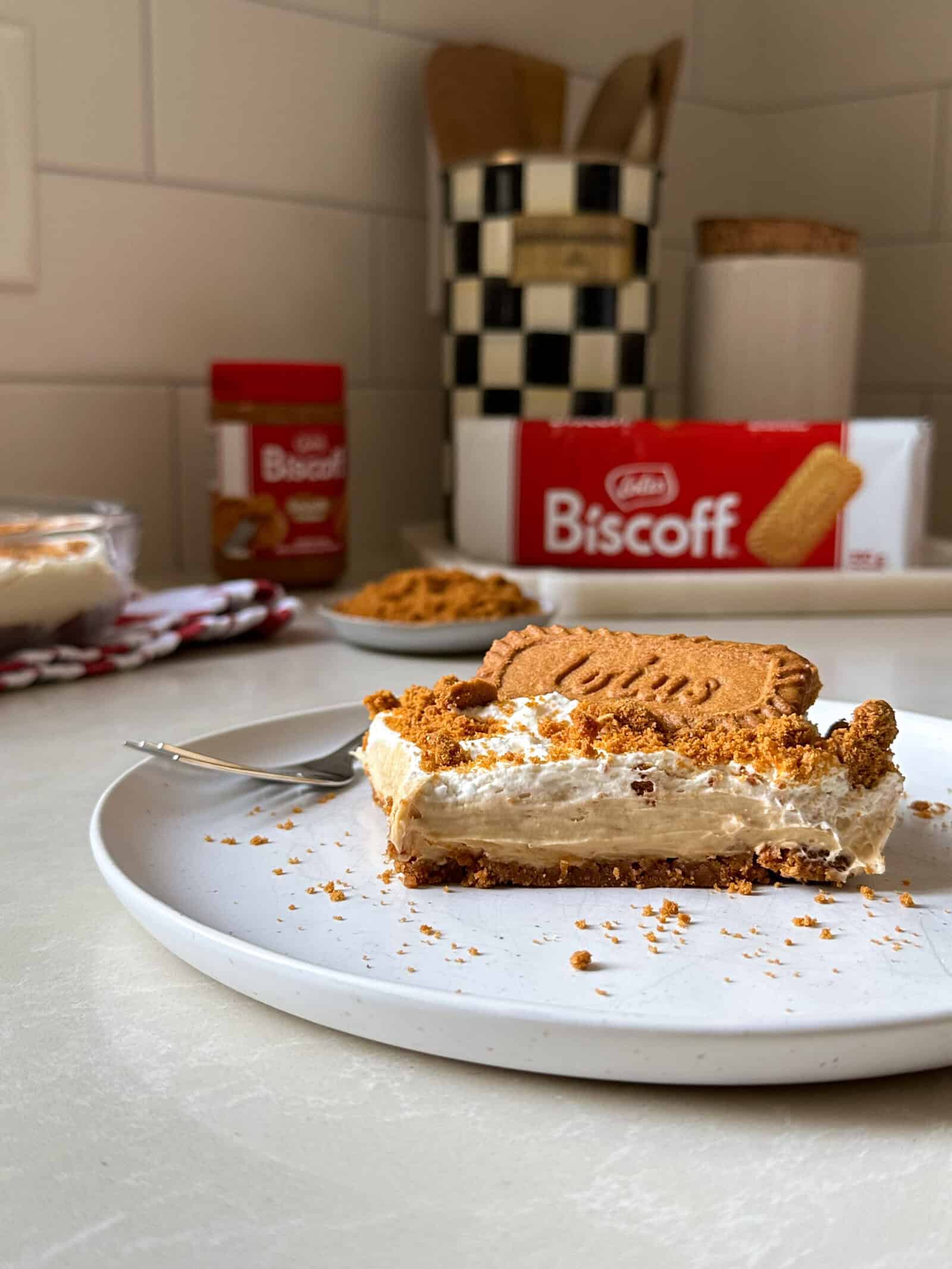 A slice of Biscoff No Bake Cheesecake topped with cookie and crumbs sits on a white plate with a fork. In the background are a Biscoff cookie box, a jar of spread, and a kitchen utensil holder, ready for your next No Bake Cheesecake Recipes adventure.