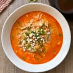 A bowl of hearty red soup with shredded meat, green onions, and white beans on a wooden surface. The soup appears rich and flavorful, garnished with green onions. A textured cloth and a dark object are partially visible beside the bowl.