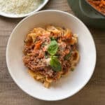 A bowl of pasta topped with a rich meat sauce and garnished with fresh basil and grated cheese. The dish is placed on a wooden table next to a small bowl of grated cheese and a green pot filled with more sauce.