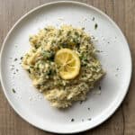 A white plate with creamy pasta garnished with chopped parsley, grated cheese, and topped with a lemon slice, set on a wooden surface.