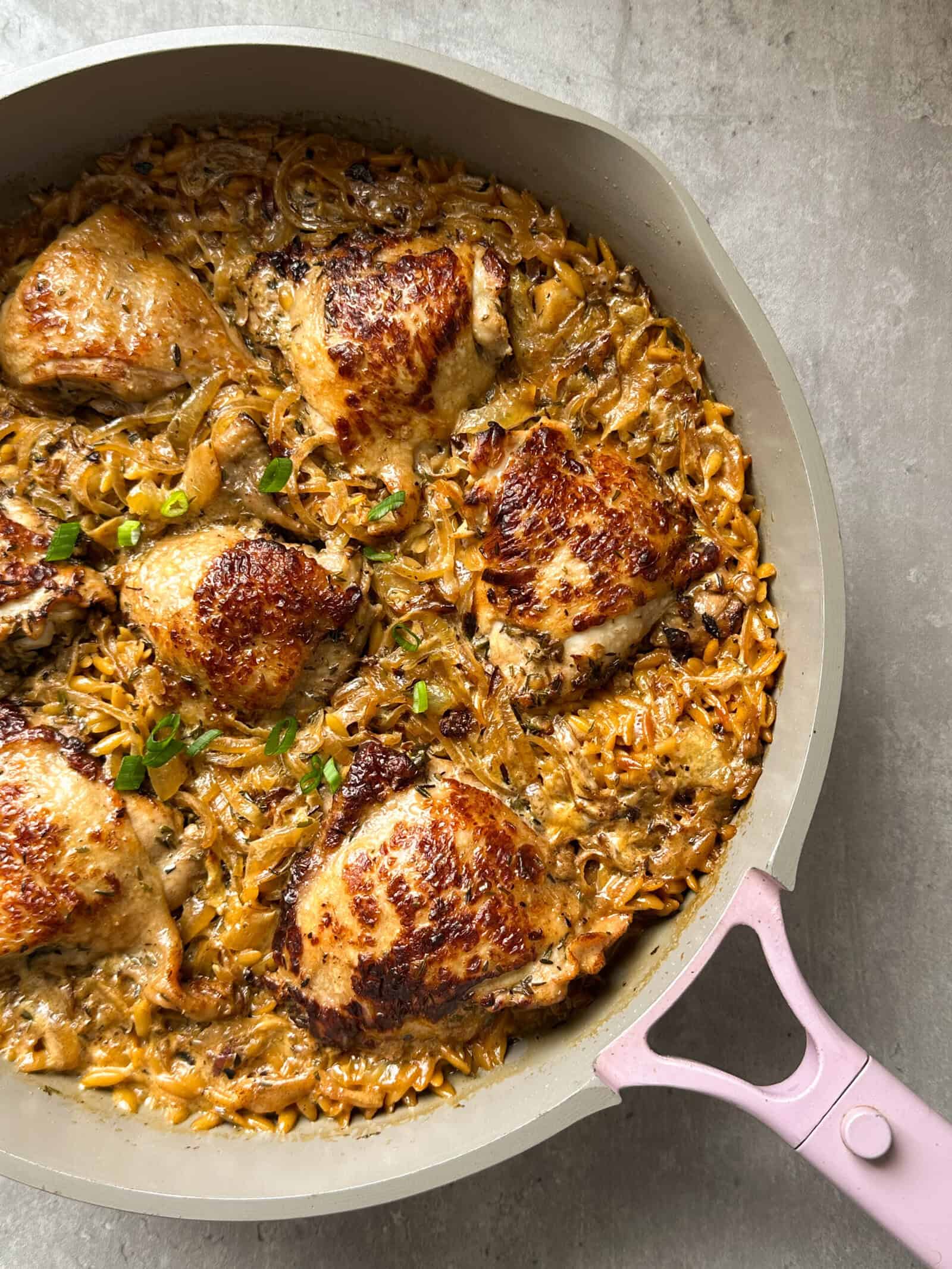 A skillet with crispy, browned chicken thighs atop a creamy orzo pasta mixture evokes the rich flavors of French Onion Chicken. The dish is garnished with fresh herbs and rests on a gray surface.