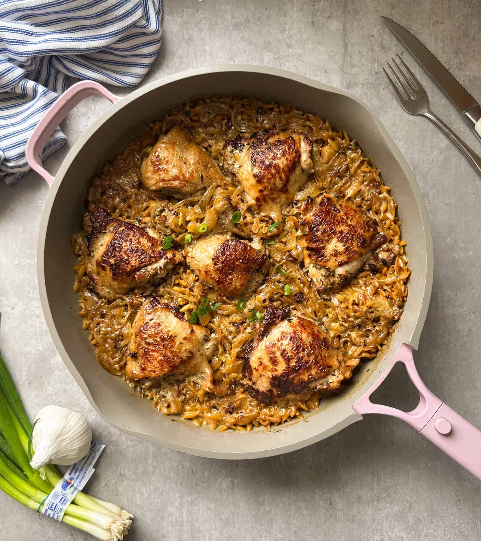 A skillet with succulent French Onion Chicken thighs on a bed of creamy rice. The dish is garnished with fresh herbs. Next to the skillet are a striped cloth, fresh garlic, and green onions. A fork and knife rest nearby on a gray surface, completing this elegant French Onion Chicken recipe tableau.