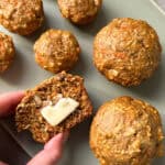 Hand holding a halved Morning Glory Muffin with a pat of butter, revealing its moist, dense texture bursting with walnuts and carrot shreds. Several golden-brown muffins rest on a green plate, showcasing the allure of this healthy muffin delight.