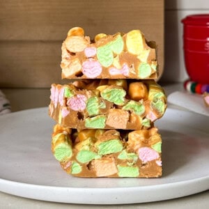 Three squares of colorful peanut butter marshmallow squares are stacked on a white plate. This easy no bake dessert features pastel mini marshmallows and peanut butter fudge, set against a neutral kitchen background.