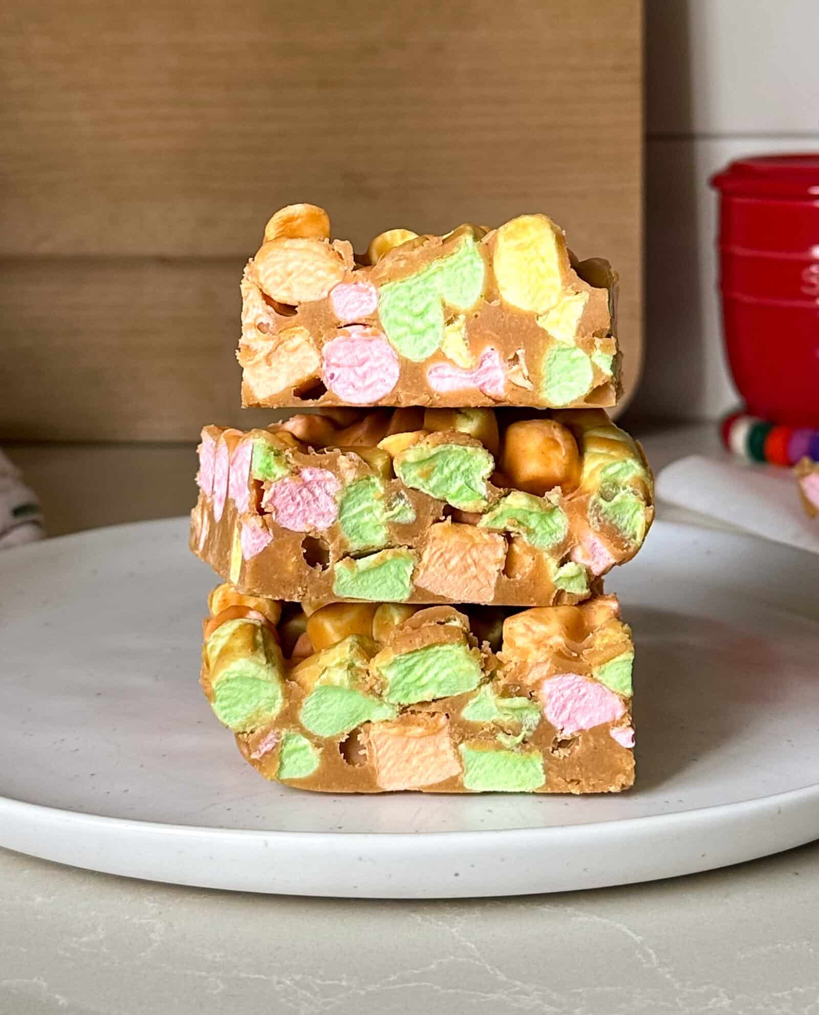 Three squares of colorful peanut butter marshmallow squares are stacked on a white plate. This easy no bake dessert features pastel mini marshmallows and peanut butter fudge, set against a neutral kitchen background.